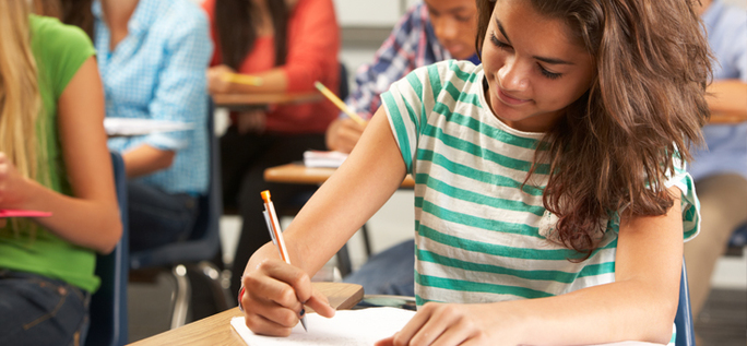 Imagem mostra menina de camiseta listrada branco e verde em uma sala de aula, escrevendo em um caderno.