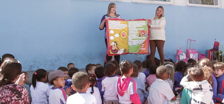 Imagem mostra duas professoras apresentando um livro colorido aos alunos