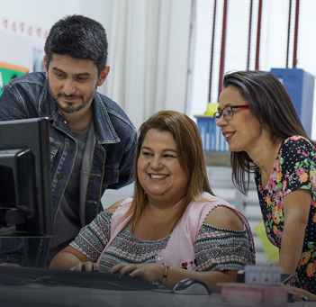 Professora em frente ao computador sorri. Ao seu lado um homem e uma mulher também olham para o computador.