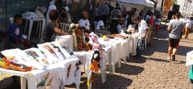 Expositores da feira afro meninas mahin, apoiados pelo Fundo Zona Leste Sustentável. Na foto, há várias mesas de plástico perfiladas, com produtos como camisetas e bonecas na temática afrobrasileira