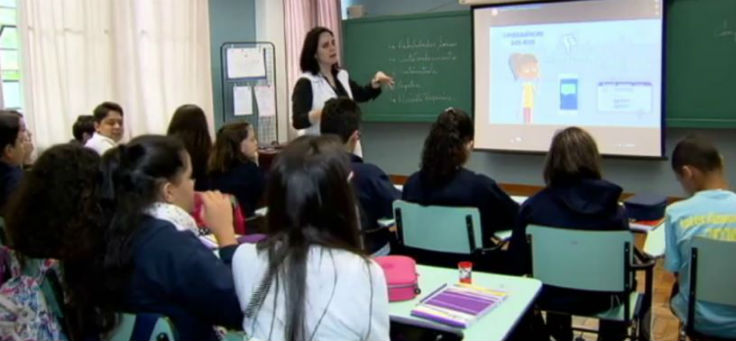 Cerca de 10 alunos sentados em carteiras, de costas, observam projeção em telão dentro da sala, enquanto professora, de pé, explica dinâmica