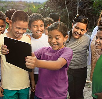 Em meio à natureza, quatro crianças sorriem encantadas, acompanham conteúdo em um tablet