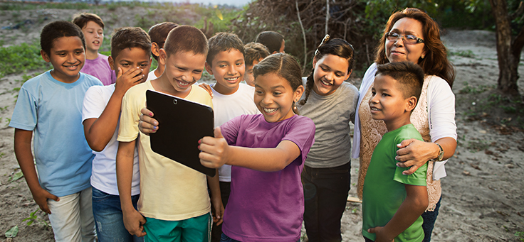 Em meio à natureza, quatro crianças sorriem encantadas, acompanham conteúdo em um tablet