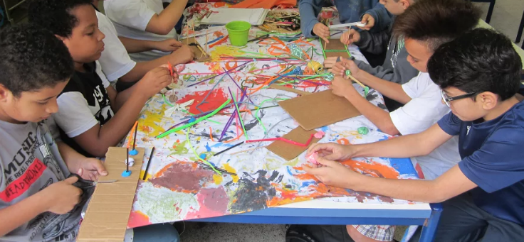 Crianças sentadas em mesa cumprida fazem pintura bem colorida sobre papel