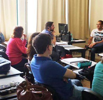 Professor está sentado em frente à lousa diante de sala cheia de alunos