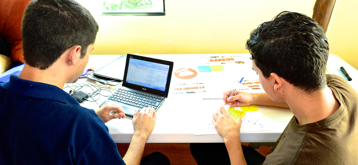 Dois jovens de costas sentados em uma cadeira apoiados em uma mesa, um com um net book, outro com uma caneta desenhando sobre uma cartolina