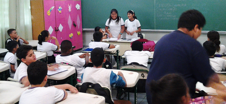 Foto mostra sala de aula a partir dos fundos, com alunos sentados em carteiras, de costas, e duas alunas de origem boliviana de pé, na mesa onde fica a professora, de costas pra lousa e de frente pros colegas, durante apresentação