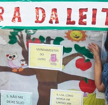 Professora está em frente a mural onde se lê “Hora da Leitura”