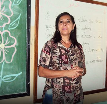 Professora está em frente a lousa onde há uma flor desenhada