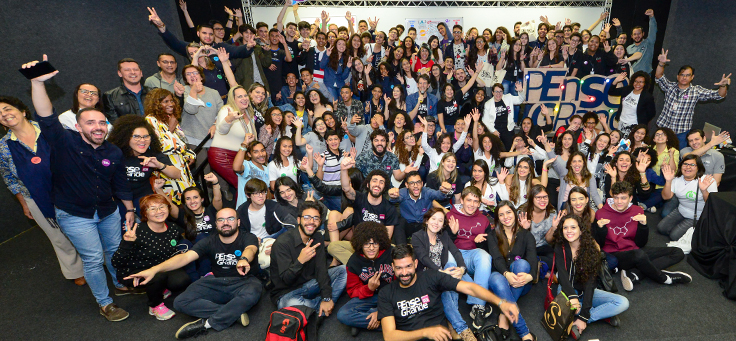 Na imagem, todos os participantes do Demoday, entre alunos, educadores, familiares e organizadores, aparecem de braços abertos, em volta de um totem com logo do programa Pense Grande