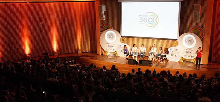 Na imagem, seis palestrantes estão sentados em roda em cima do palco durante o evento Educação 360