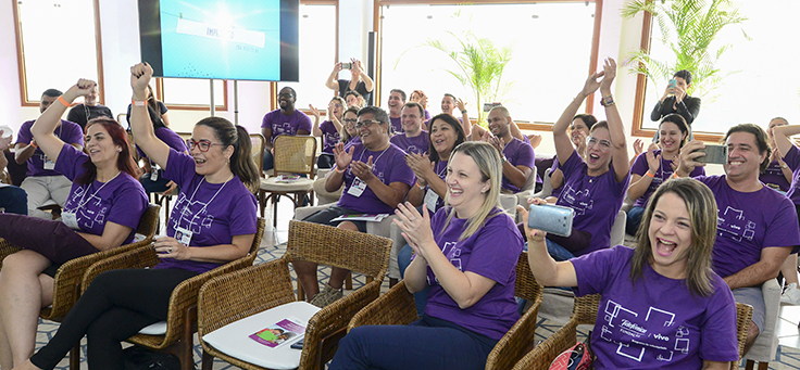 Na imagem, colaboradores do programa de voluntariado usam camiseta púrpura do programa. Eles estão sentados em uma plateia, aplaudindo e comemorando durante evento