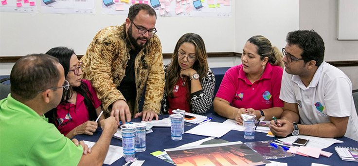 Beto Silva, que conduz oficina sobre futurismo, está em pé orientando cinco educadores sentados em uma mesa