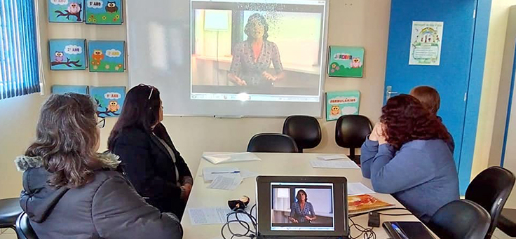 Quatro educadores sentados em volta de uma mesa na escola, assistem a uma projeção de vídeo como parte da formação do Aula Digital nas escolas de Viamão