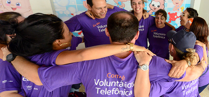 Colaboradores da Telefônica estão se abraçando, formando um círculo. Eles usam camiseta púrpura na qual se lê: Comitê Voluntários Telefônica