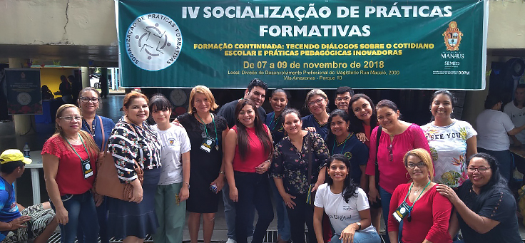 Grupo formado por cerca de 10 professores e participantes em geral posa embaixo de banner do evento de boas práticas Socialização de Práticas Formativas