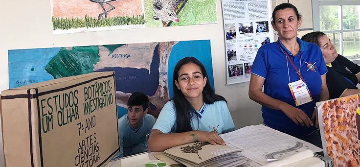 Aluna está sentada ao lado de cartaz em que se lê Estudos Botânicos, um olhar investigativo, durante atividade em mostra de boas práticas pedagógicas realizada em Viamão-RS.