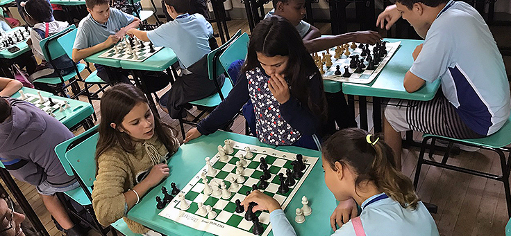 Em primeiro plano, duas jovens disputam partida de xadrez e uma terceira as observa em pé durante atividade em mostra de boas práticas pedagógicas realizada em Viamão-RS.