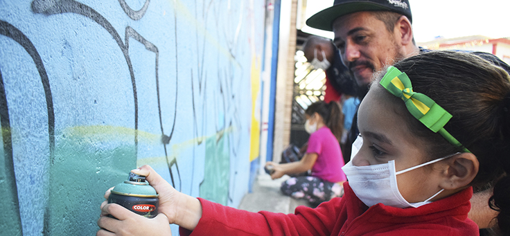 Criança usa spray para pintar mural e é observada por instrutor do Cidade na Escola, que promove a educação integral