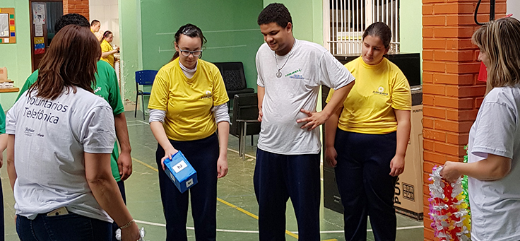 Na imagem, grupo de alunos da Escola Primavera estão em pé diante de uma espécie de tabuleiro desenhado com fita no chão durante dinâmica sobre programação