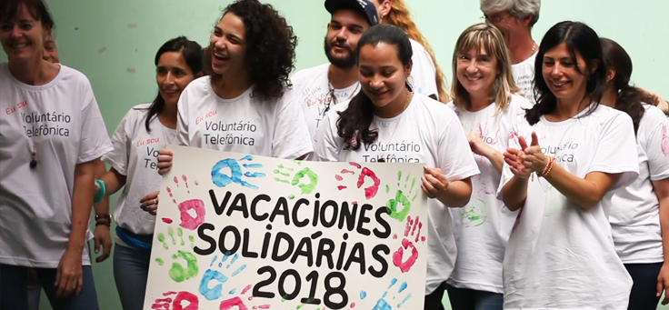 Na imagem, colaboradores vestindo a camiseta do programa Vacaciones Solidárias seguram cartaz ilustrado com carimbo das mãos de jovens da Escola Especializada Primavera.