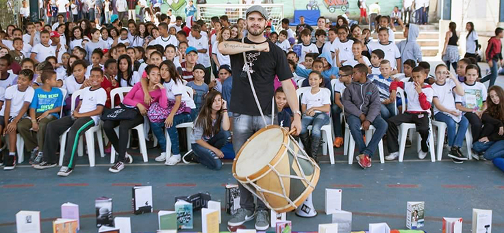 O professor Rodrigo Ciríaco, criador do Sarau dos Mesquiteiros ao lado de alunos, segura um tambor. Ao fundo é possível ver uma plateia formada por jovens