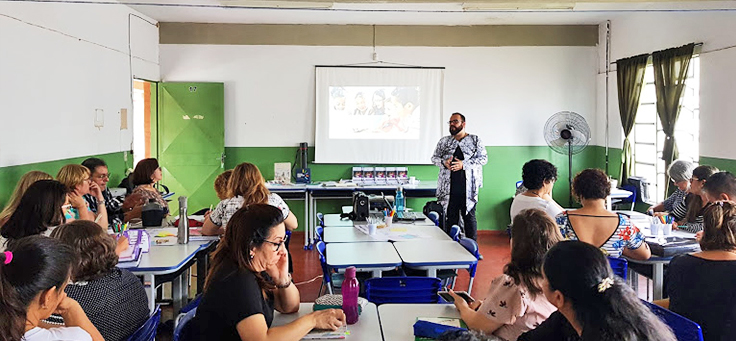 Beto Silva fala para professores sentados em grupos durante palestra formativa da Assessoria Plugada, iniciativa da Fundação Telefônica Vivo que atua para fortalecer a inovação educativa em redes públicas.