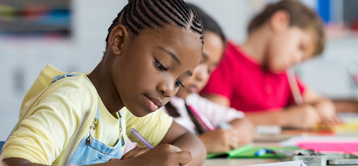 Menino está sentado em uma carteira escrevendo em um caderno. Imagem ilustra texto sobre pensadores que embasam o conceito de inovação em educação.