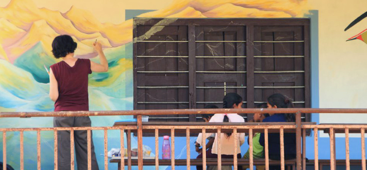 Mulher desenha quadro em muro de escola enquanto duas meninas conversam sentadas em banco atrás dela – matéria sobre volunturismo foi destaque em Voluntariado em 2018.