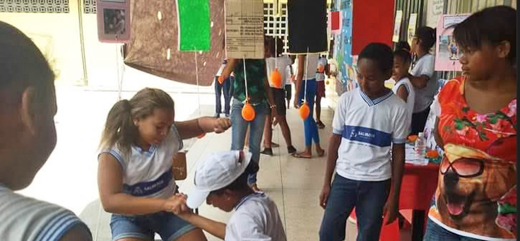 Duas alunas da Escola Municipal Gersino Coelho estão agachadas e de mãos dadas em demonstração de brincadeiras que resgatam culturas marginalizadas, no projeto É de Quebrada que Eu Vou.