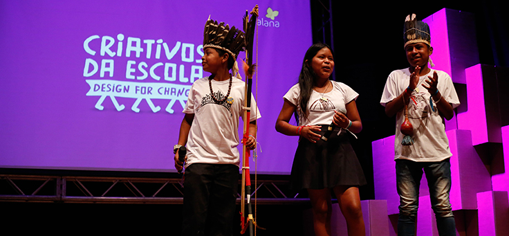 Três alunos, usando cocar e arco e flecha, estão no palco do prêmio Criativos da Escola, onde o dicionário das línguas ofaié e guarani ganhou prêmio ao resgatar culturas marginalizadas.