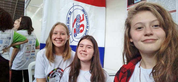 As alunas Manoela Kieling, Laura Schuster e Julia Dapper, que ajudaram a desenvolver o aplicativo de combate ao bullying, estão sorrindo para a foto com uma bandeira da Escola Municipal de Ensino Fundamental 25 de Julho ao fundo.