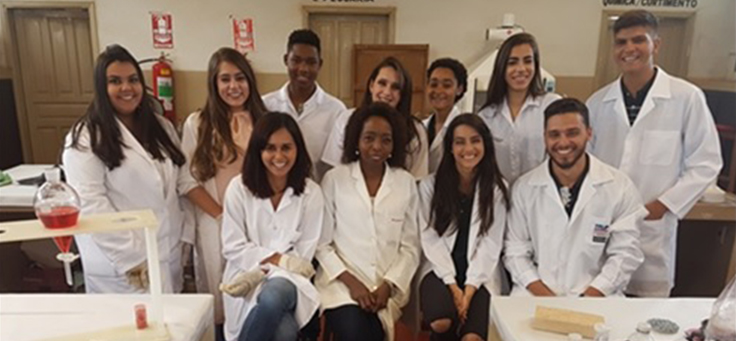 Alunos da professora Joana, posam para foto em grupo, vestidos de jaleco dentro do laboratório.