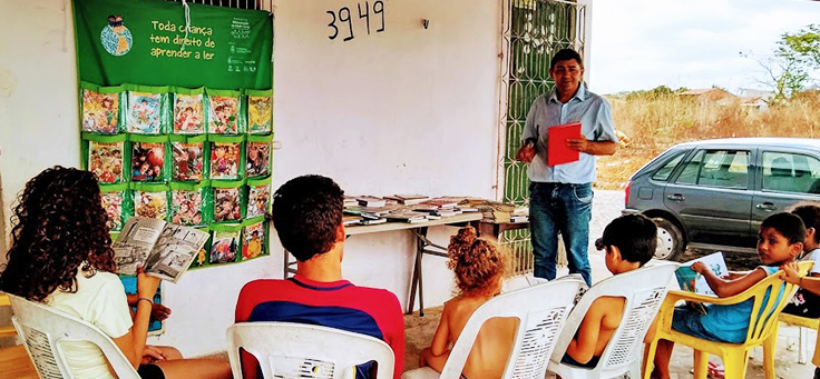 O professor José Gilson Lopes, que desenvolveu o projeto Mala de Leitura para incentivar a inclusão por meio do hábito de ler, está em pé em frente a alunos, mostrando uma mesa cheia de livros.