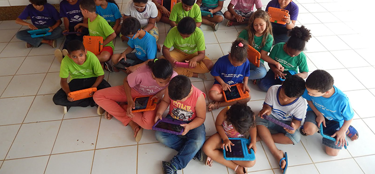 A imagem mostra crianças sentadas no chão com tablets nas mãos.