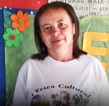 A professora Cleide posa para a foto atrás de um cartaz e usa uma camiseta que diz: Feira Cultural