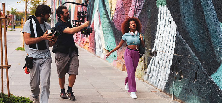 Na foto, a apresentadora Lellêzinha aparece caminhando na frente de um muro grafitado. Ela está sendo filmada por uma equipe de gravação