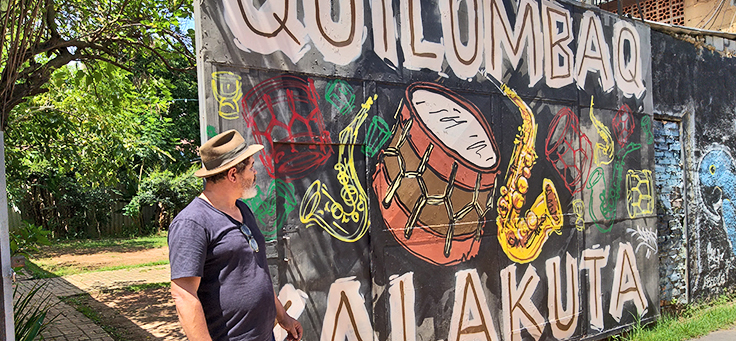 O arte-educador e gestor José Soró está olhando para o portão da sede da Comunidade Cultural Quilombaque em Perus, que tem instrumentos de sopro e um tambor desenhados.