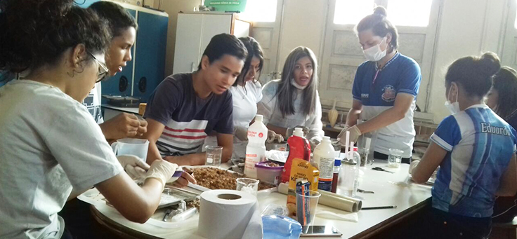 Grupo de alunos está sentado em torno de uma bancada com elementos usados para experimento, como álcool e alimentos, em laboratório da Escola Estadual Nossa Senhora de Nazaré, em Manacapuru (AM).