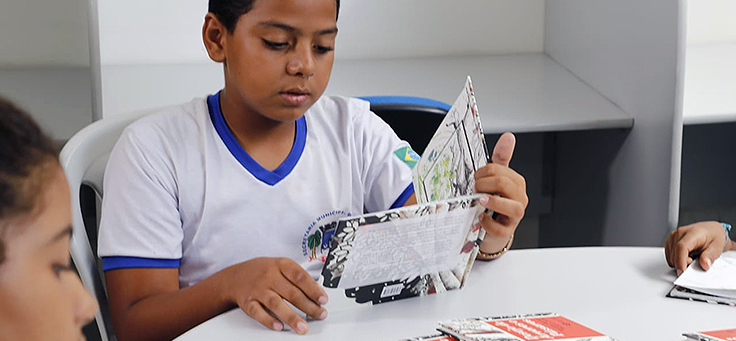 Menino vestindo uniforme escolar está sentando atrás de uma mesa lendo um livro