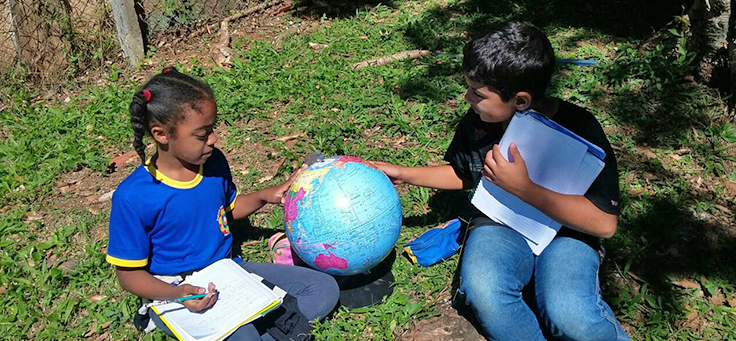 Duas meninas da Escola Municipal Professor Ivan Galvão de França, que integra lista das mais inovadoras do Brasil, estão sentadas lado a lado manipulando um globo terrestre.
