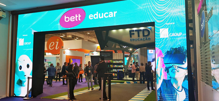 A imagem mostra a entrada do evento Bett Educar, com uma fachada feita com um painel de led, e algumas pessoas ao fundo da foto.