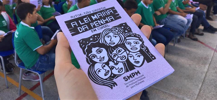 Em primeiro plano uma mão segura e mostra a capa da versão em cordel, onde estão ilustradas sete mulheres de várias etnias, de obra que narra a Lei Maria da Penha para jovens de escolas no Ceará.