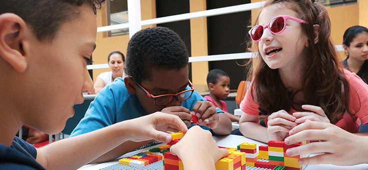 Dois meninos e uma menina estão interagindo em uma bancada com peças do projeto Lego Braille Bricks, que promove a alfabetização em braile. Um dos meninos está empilhando peças enquanto conversa com a garota, que usa óculos rosa.