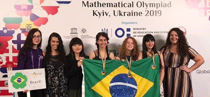 Mariana Groff, Ana Beatriz Studart e Maria Clara Werneck estão segurando bandeira do Brasil e com medalhas de ouro e bronze no pescoço no pódio da 8ª Olimpíada Europeia Feminina de Matemática. Elas incentivam a presença de meninas na Matemática.