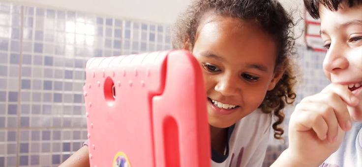 Imagem mostra duas crianças olhando para um tablete e sorrindo