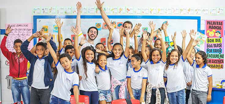 Alunos da Escola Municipal de Ensino Fundamental Frei Fernando estão com braços levantados, posando ao lado de dois participante do programa de voluntariado do Vacaciones Solidárias.