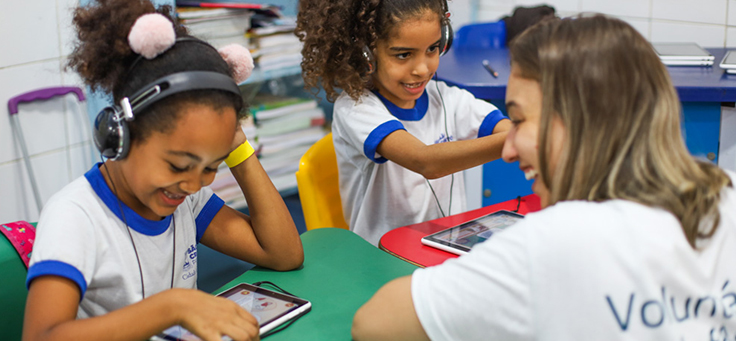 Voluntariado em escolas é uma opção para aproximar a comunidade do ambiente escolar. Na imagem, voluntária interage com estudante que está navegando em tablet e usando fones de ouvido.