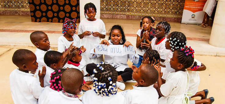 Na imagem, professora está rodeada de alunos e segurando cartaz em que se lê algumas sílabas. Em Angola programa usa atividades lúdicas para inovar ensino-aprendizagem.