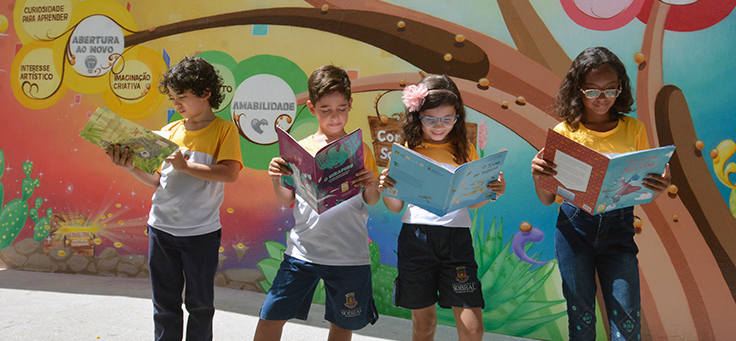 Quatro crianças estão segurando livros em frente a um muro com desenhos coloridos em escola de Sobral (CE), que tem rede de ensino modelo em alfabetização de qualidade.